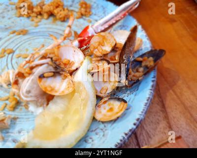 Leftover seafood paella featuring clams, mussels, norway lobster, rice, and a slice of lemon rests on a textured, light blue plate on a wooden table, Stock Photo