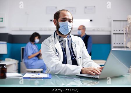Caucasian medic specialist using laptop for patient appointments and looking at camera. Portrait of young doctor with face mask checking emails and medical records on digital device in clinic office. Stock Photo