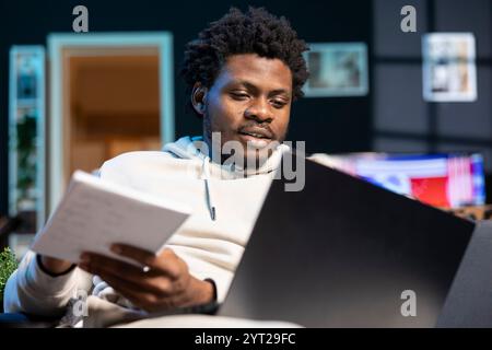 Freelancer working from dimly lit living room, writing startup ideas on paper. African american man feeling thrilled after coming up with entrepreneurship plan, using pen to note it down Stock Photo
