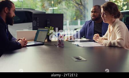 Dealership salesman looking over appointment list on laptop spreadsheet to schedule car test drive for customers. Showroom agent discussing with clients, organizing test drive, camera B Stock Photo