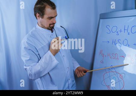 A scientist in a lab coat and glasses pointing at chemical formulas on a whiteboard, emphasizing science education, research, and explanation of compl Stock Photo