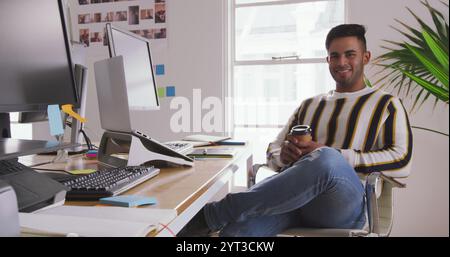 Portrait of a happy biracial man working in a creative office, using graphic tablet and laptop compu Stock Photo