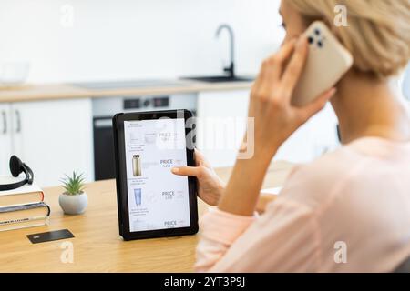 Caucasian woman using tablet to browse cosmetics online while talking on phone. She is ordering products from delivery website at kitchen counter. Stock Photo