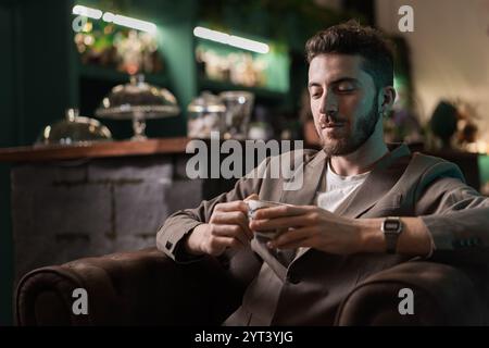 Stylish young man in beige blazer enjoying afternoon tea in atmospheric cafe. Modern professional taking mindful break in luxury setting with moody gr Stock Photo