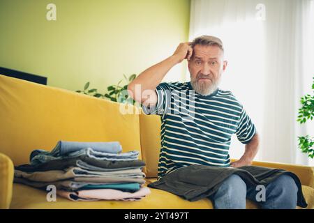 Elderly bearded man in casual wear confused while organizing clothes on yellow couch in bright living room Stock Photo
