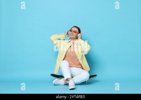 Smiling woman with headphones sitting on skateboard against light blue background Stock Photo