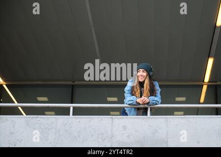 Young woman wearing headphones leaning on railing and smiling outdoors Stock Photo