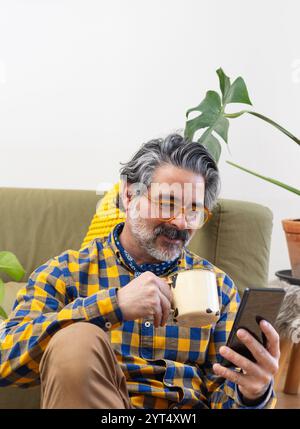 Man using a smartphone sitting in his living room drinking coffee Stock Photo