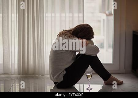 Lonely woman sitting on floor by the window with glass of wine Stock Photo
