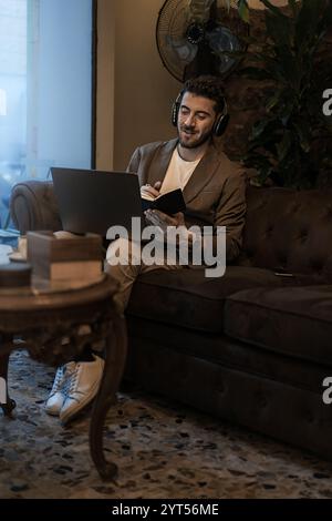 Modern businessman wearing headphones working on laptop and taking notes in stylish cafe. Young professional multitasking in comfortable workspace wit Stock Photo