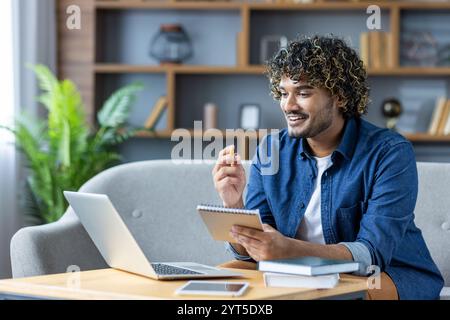 A young man is engaged in online learning, sitting on a comfortable sofa with his laptop and a notebook in hand. He appears focused and enthusiastic, using digital tools for his studies. Stock Photo
