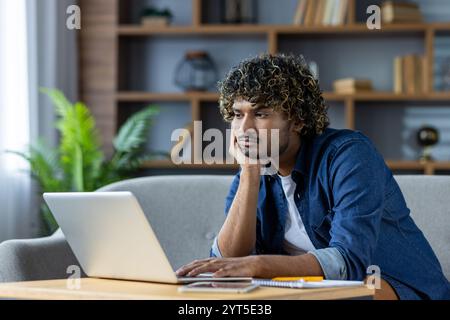 A young man with curly hair appears bored while using a laptop at home. He rests his head in his hand, illustrating a common work-from-home experience. This image captures feelings of monotony Stock Photo