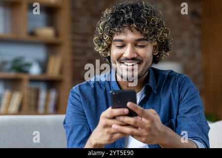 A cheerful man enjoys browsing on his smartphone in a comfortable living room setting. His positive expression suggests a connection with friends or engaging content. T Stock Photo