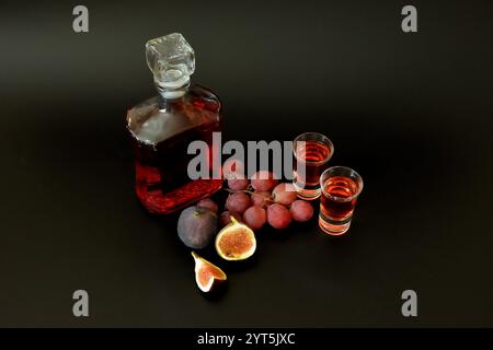 Homemade fruit liqueur made from figs and red grapes on a black background, in a bottle and two glass goblets. Close-up. Stock Photo