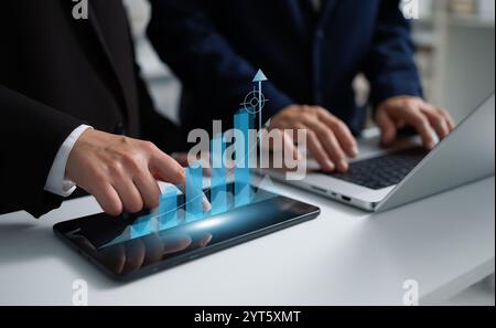 A businessman and businesswoman collaborate over a laptop, while the businesswoman points at a tablet displaying a holographic growth chart. A powerfu Stock Photo