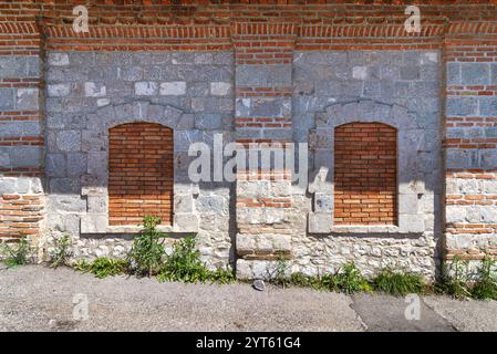 Brick wall with two arched windows. The wall is made of a combination of red bricks and grey stones. The windows have brick frames Stock Photo