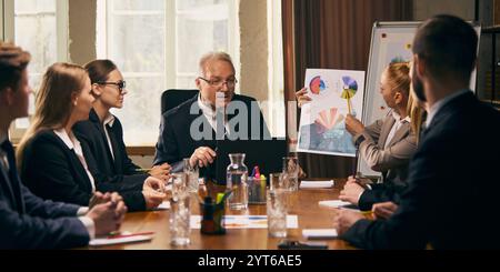 Business team closely analyzing charts and data presented by female colleague during formal meeting. Senior Leader attentively listening. Stock Photo