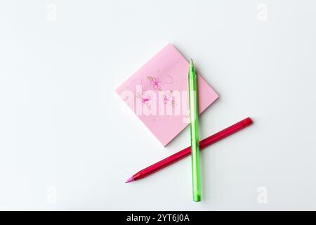 Pink and green color ballpens beside a square sticky notepad with doodle flowers drawn on it. Creative stationery for art lesson. Stock Photo