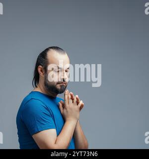 Studio shot on a gray background depicting a pensive adult man with clasped hands, looking down in reflection, conveying emotions, feelings, thoughts, Stock Photo