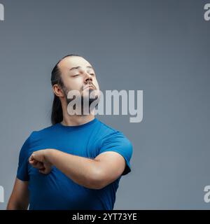 Arrogant young man with a beard and long hair, wearing a blue t-shirt, beating his chest like tarzan, exuding pride and self-importance against a gray Stock Photo