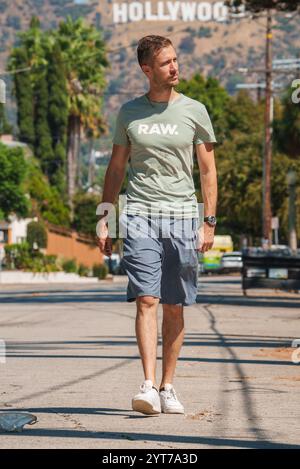 A man in a light green T shirt and shorts walks down a palm lined street in Los Angeles, California, with the iconic Hollywood Sign visible in the bac Stock Photo