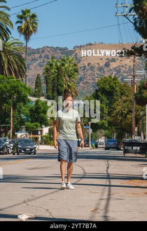 A man in a light green T shirt and shorts walks down a palm lined street in Los Angeles. The iconic Hollywood Sign is visible under a clear blue sky. Stock Photo