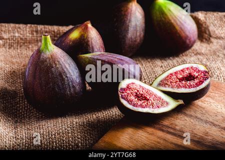 Ripe figs displayed on a rustic burlap cloth, with one fig cut open to reveal its vibrant red flesh and seeds, resting on a wooden surface under soft Stock Photo