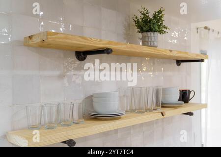 Two wood shelves with industrial pipe brackets on a white tile wall. Dishes, glasses, and a potted plant are displayed. Stock Photo