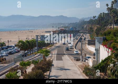 View of Pacific Coast Highway in Santa Monica, California, with beach and ocean on the left, bustling traffic, and palm trees lining the road. Stock Photo