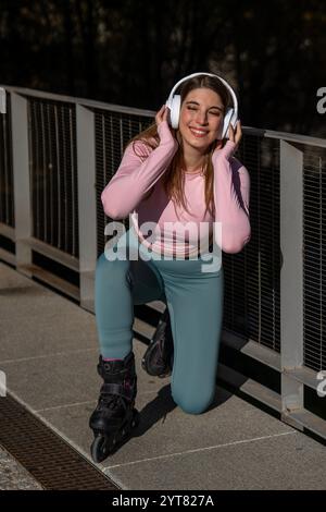 Young sporty woman wearing inline skates and headphones is kneeling and smiling Stock Photo
