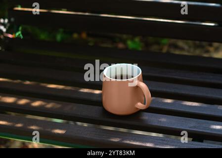A coffee cup sits on a wooden bench in the shade. The cup is half full of coffee. Concept of relaxation and leisure. Mocha Mousse color of the year 20 Stock Photo