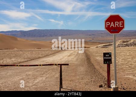 Desert road blocked by a 'Pare' stop sign and 'No Pasar' warning in the Valle de La Luna, Chile, with sand dunes and mountains in the background Stock Photo