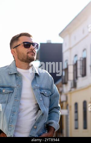 Caucasian urban man in sunglasses and denim jacket walking on city street. Stock Photo