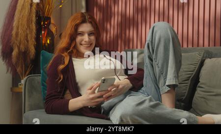 Smiling woman female homeowner Caucasian girl tenant customer lying on couch sofa in apartment happy ginger lady user addict mobile phone using Stock Photo