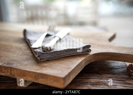 Simple rustic table setting on wooden cutting board. Old silver cutlery with linen napkin and black ceramic dishes. Stock Photo