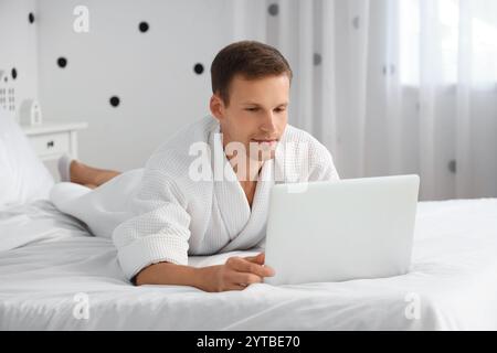 Young man in bathrobe using laptop on bed at home Stock Photo