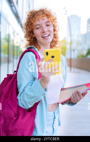 Smiling young woman holding smartphone and notebooks outdoors in casual attire Stock Photo