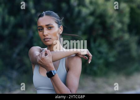 Slim woman stretching her arm and looking at camera. Confident sportswoman warming up a hand before an outdoor workout. Stock Photo