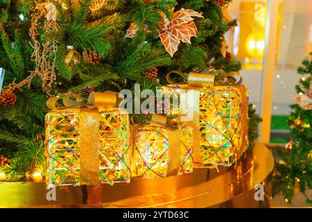 Christmas tree decorated with yellow gold balls and sparkling lights installed indoors, boxes gifts with gold satin silk ribbon for Christmas and New Stock Photo