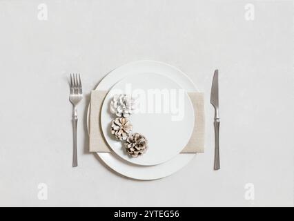 Table setting with silverware, napkin, cones and a white plate on a white background. Christmas or New Year dinner. Copy space. Top view, flat lay. Stock Photo