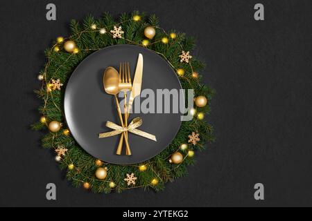 Black plate in a Christmas wreath with gold cutlery and decor on a black background. Festive table setting. Copy space. Top view, flat lay. Stock Photo