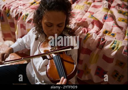 A focused woman plays violin in a warm, cozy room featuring a vibrant patchwork quilt, creating a serene, musical atmosphere. Stock Photo