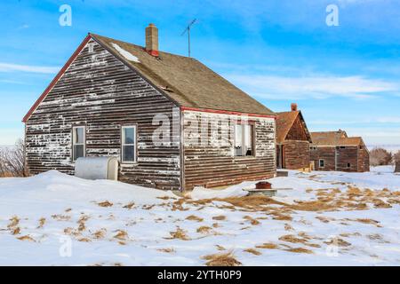 A small, old house with a chimney sits in a snowy field. The house is surrounded by other small houses, and the sky is clear and blue Stock Photo