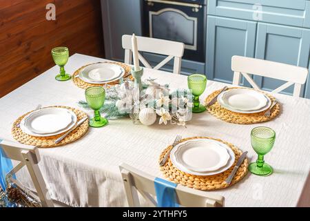 Holiday dining table is elegantly arranged with green glassware, woven placemats and white plates, creating warm and inviting setting on modern kitche Stock Photo