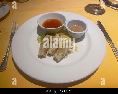 A white plate of three Thai Spring Rolls; deep-fried with minced shrimp, vegetables, peanuts, and sweet chili sauce. Silverware surrounds the dish, al Stock Photo