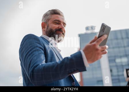 A happy mature business man using a mobile phone outside with urban buildings in the background.  Stock Photo