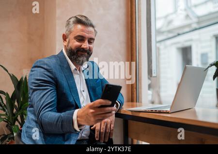 A happy mature business man using his phone while working on a laptop in the office or cafe restaurant. Stock Photo