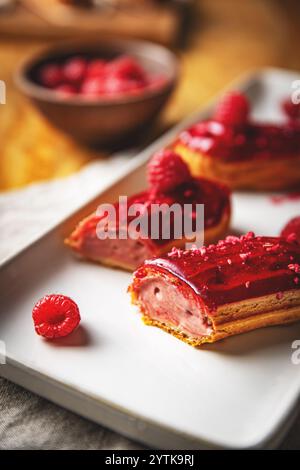 Freshly made raspberry eclairs are topped with red icing and raspberry bits, served on a white plate Stock Photo