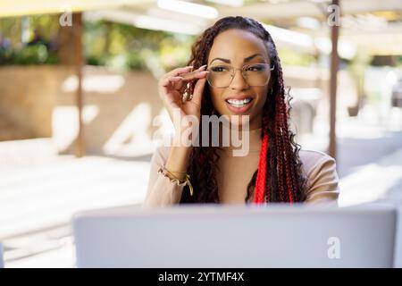 A cheerful woman in glasses sits at a laptop outdoors, smiling as she works on tasks Stock Photo