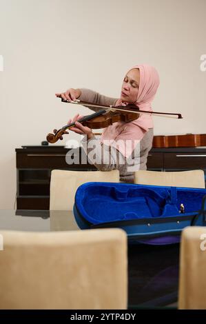 A woman stands playing the violin in a home setting, expressing deep concentration and emotion, with an empty violin case on the table nearby. Stock Photo